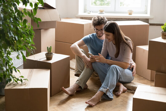Happy couple sitting on floor among moving boxes using phone - Powered by Adobe
