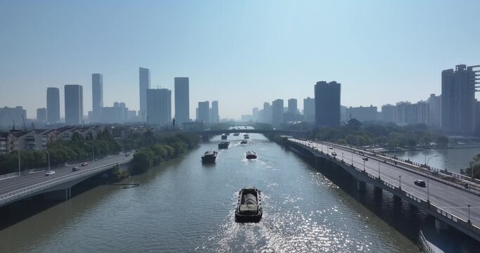 Cargo boats navigate a city river flanked by modern high-rise buildings and bridges under clear daylight
