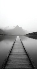 Fototapeta premium Misty lake vista with wooden pier. A tranquil grayscale image of a wooden pier extending into a calm lake. Foggy mountains rise in the distance, mirrored in the water's surface.