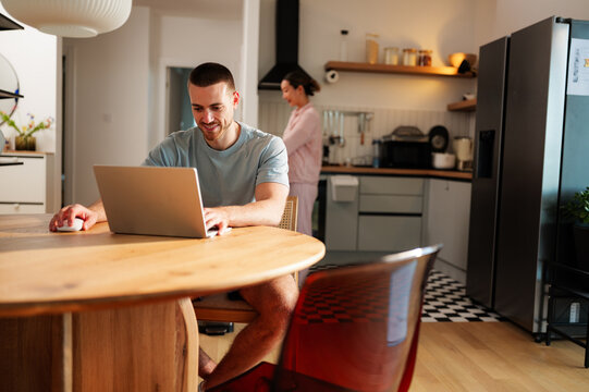 Young man smiling and working on his laptop at the dining table while his partner prepares breakfast in the kitchen, enjoying the benefits of remote work and flexible schedules