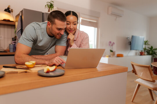 Happy couple using laptop in modern kitchen, browsing internet and enjoying time together, with fresh apples on the counter indicating a healthy lifestyle
