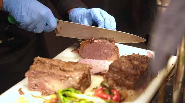 Close up of a chef's hands in blue gloves cutting a medium-rare roast beef on a cutting board.
