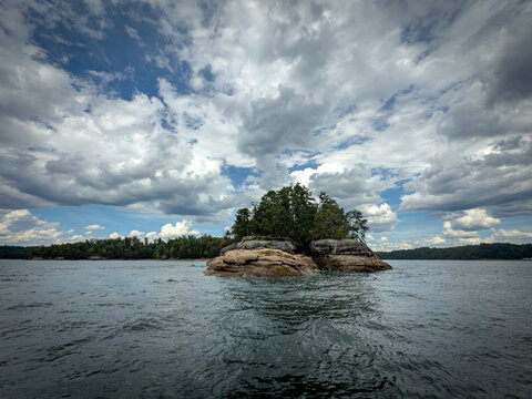 Rocky sandstone outcrop with dense trees surrounded by the waters of Laurel River Lake, Kentucky. A peaceful wilderness scene with textured rock formations, lush forest, and striking cloudy skies.