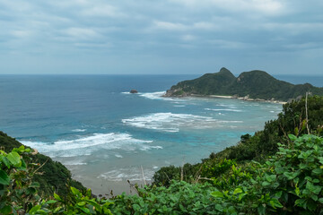 Powerful white-capped waves relentlessly crash against a rugged, rocky coastline below a green...