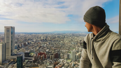 A young man wearing a grey beanie and hoodie looks out from a tall skyscraper observatory,...
