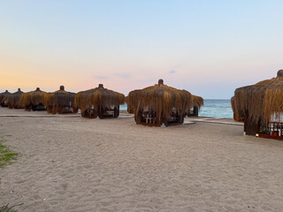 Thatched cabanas line the sandy beach under the pastel evening sky. Coastal leisure, tourism and...