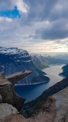 A person sits peacefully in contemplation on the edge of the famous Trolltunga rock, enjoying the serene and breathtaking sunset view over the Ringedalsvatnet lake and snow-covered mountains.