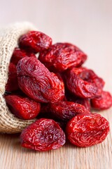 A burlap sack overflows with vividly colored dried red fruits arranged on a wooden surface. These...