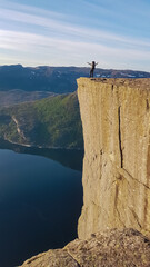 A lone hiker stands with arms open wide on the iconic Preikestolen cliff during a golden sunset,...