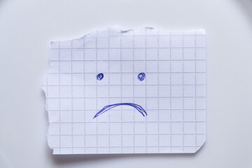 A sheet of squared paper with a sad face drawn in blue pen lies on a white background as a symbol...