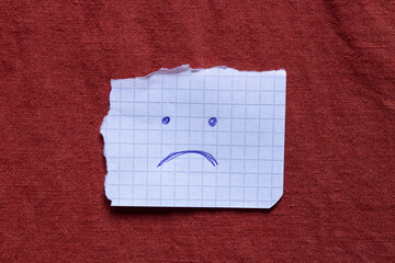 A piece of squared paper with a sad face drawn with a blue pen lies on a red cloth as a symbol of...