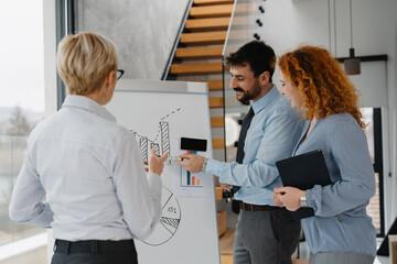 Business team during a presentation in the office. A woman is pointing at a flipchart with bar and pie charts, while two colleagues listen attentively and take notes.
