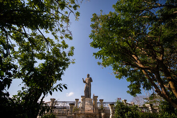 A large stone statue stands elevated between two lush trees, with a stone fence and a clear blue...