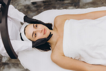 Beautician examining face of young woman lying on massage table
