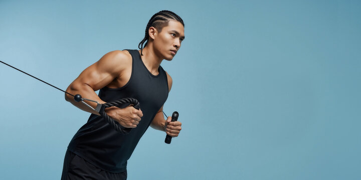 Muscular Asian man doing a strength training exercise on a cable machine. Focused athlete working out against a blue background. Copy space. - Powered by Adobe