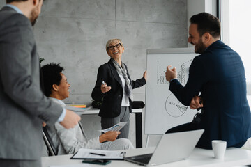 Group of cheerful business professionals standing together in the office. They are dressed in formal business attire, smiling, and posing with arms around each other
