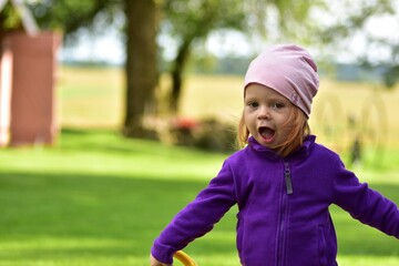 Fototapeta premium Smiling toddler girl with pink hat and purple jacket stands outside in countryside garden, opening mouth playfully. Bright childhood portrait in rural family environment.