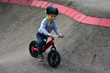 Young boy wearing helmet rides red balance bike on outdoor pump track with high curbs. Exciting...