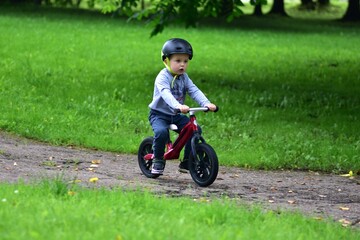 Little boy wearing safety helmet rides red balance bike on paved park path surrounded by trees and...