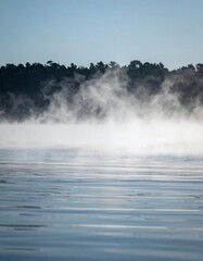 Misty Lake Landscape with Dense Fog Over Blue Water and Dark Green Trees in Background