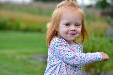 Close-up portrait of happy red-haired toddler in floral shirt outdoors. Child smiling with innocent...