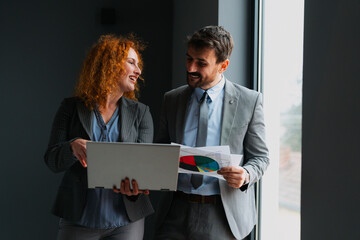 Red-haired businesswoman and businessman reviewing analysis on a laptop while holding documents in a modern office.