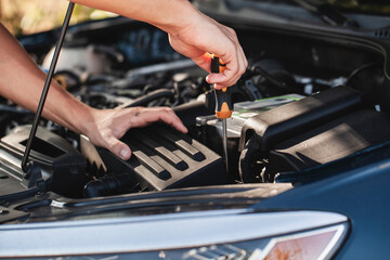 Car owner performs routine maintenance on engine using screwdriver in sunny outdoor environment