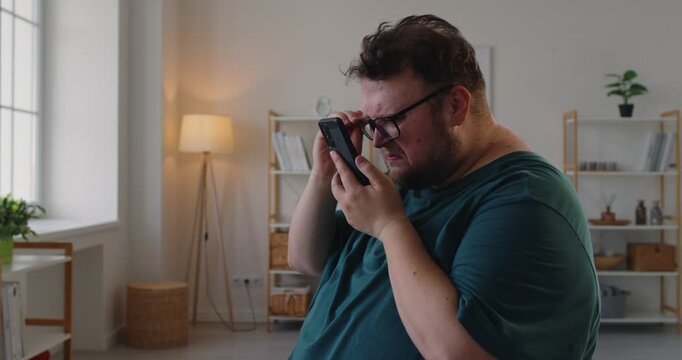 Fat overweight man with glasses holds smartphone, squinting at screen as he tries reading, struggling for focus and better vision. Man with farsightedness eye disease squints at phone screen