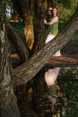 A woman in a green blouse and white trousers stands among tree trunks, her reflection visible in the water below