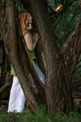 A woman in a green blouse and white trousers stands among large tree trunks, touching the bark with a calm expression
