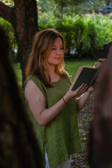 Fototapeta premium A woman in a green linen blouse reads a vintage book outdoors, framed by trees in soft evening light