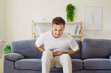 Man with stomach pain holds abdominal area. Seated on couch, he leans forward in discomfort with...
