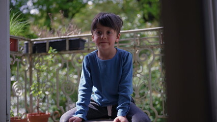 Boy sitting on a wooden stool on a balcony, surrounded by greenery, with a contemplative...