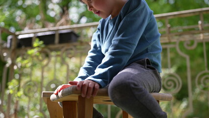 Close-up of a boy sitting on a wooden stool outdoors, hands gripping the edges, surrounded by lush...