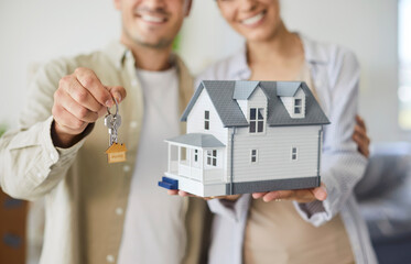 Happy couple holds house keys after purchase. They smile with a model home, marking realty success...