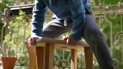 Close-up of a boy sitting on a wooden stool outdoors, hands gripping the edges, surrounded by lush...