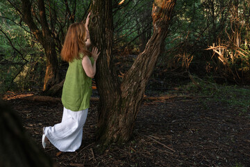 A woman in a green blouse and white trousers stands among large tree trunks, touching the bark with...