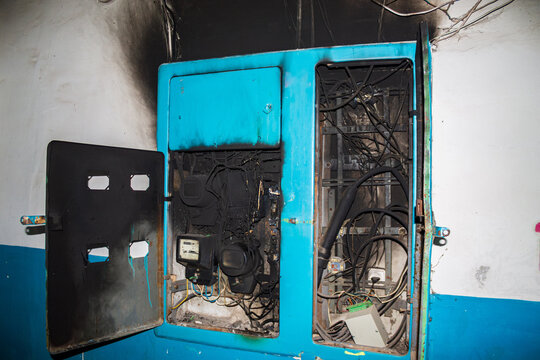 A severely burnt and damaged blue electrical panel is open, revealing scorched wires and meters. The surrounding wall also shows smoke damage, indicating an electrical fire.
