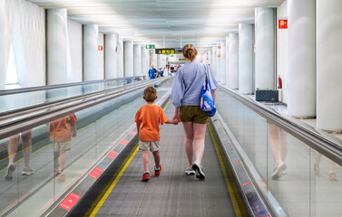 Mother and child walk hand in hand along a moving walkway with reflections and columns inside Palma de Mallorca PMI airport terminal bright interior