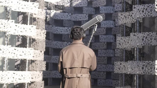 A man in a trench coat stands with his back to the camera, aiming a rifle at a patterned target wall