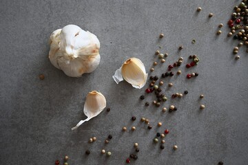 A whole garlic bulb and separated cloves are arranged with a mix of multi-colored peppercorns on a textured gray background, showcasing the raw ingredients for cooking