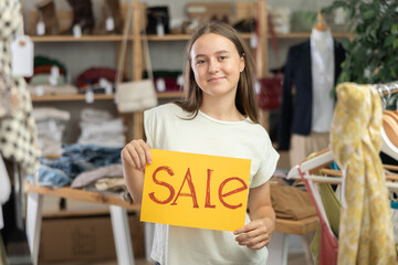 Positive teen girl standing with banner announcing summer sale in fashion boutique