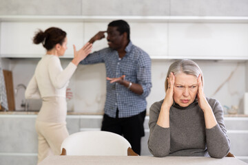 Sad old woman sitting at the kitchen table with her back to middle-aged couple quarreling together