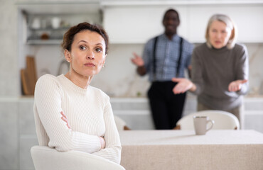 Sad middle-aged woman sitting at the kitchen table with her back to man and old woman quarreling to...