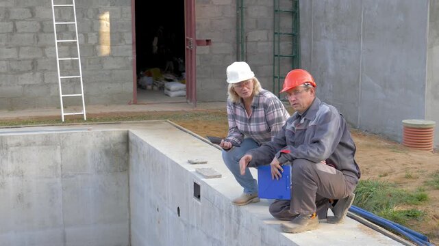 Two engineers woman and man, wearing hard hats and work clothes. Builders crouch at edge of empty unfinished concrete pool at construction site. Architect and foreman discussing plans and pointing
