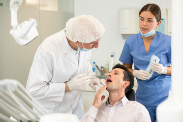 Obraz premium Man sits in armchair during visit to senior male dentist and undergoes diagnostic procedure. Doctor examines patient and asks for help of girl nurse, gives instructions on how to prepare instrument