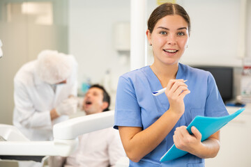 Young woman nurse in blue suit uniform with paper is standing in dental office, clinic employee in...
