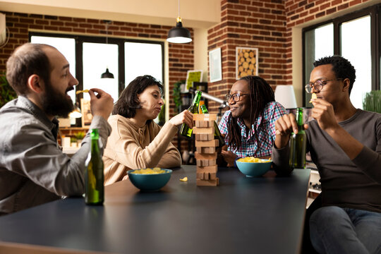 Multiethnic group of friends enjoying break from playing games, sharing snacks and drinks around table. Caucasian and african american women cheer with beer bottles as men munch on crisps. - Powered by Adobe
