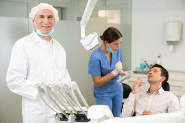 Mature male doctor is standing in dental office, clinic employee in suit uniform near dentists...