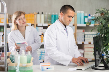 Fototapeta premium Qualified male pharmacist working at a computer against the background of a female pharmacist, they check the availability of medicines in the electronic table and in the pharmacy warehouse
